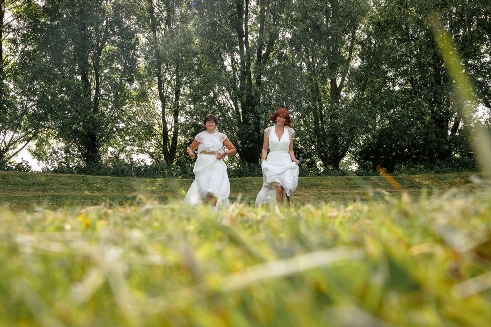 Two people in white dresses run through a sunny meadow with trees in the background on a summer day.