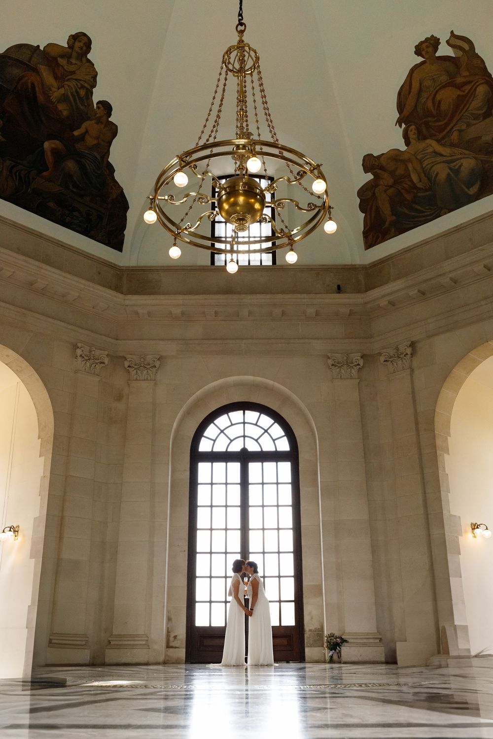 Elegant chandelier hangs in grand marble hall with tall arched window and ornate architectural details.
