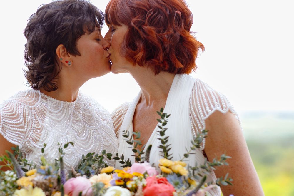 Two brides in white lace dresses kiss while holding colorful wildflower bouquets against a nature backdrop.