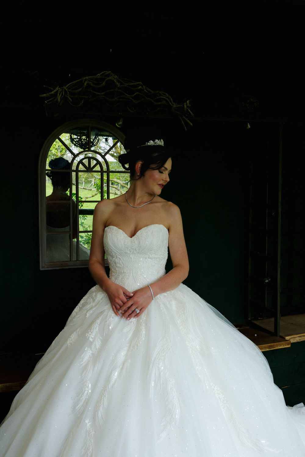 A person in an elegant white strapless ball gown wedding dress stands by an arched window in a dark room.