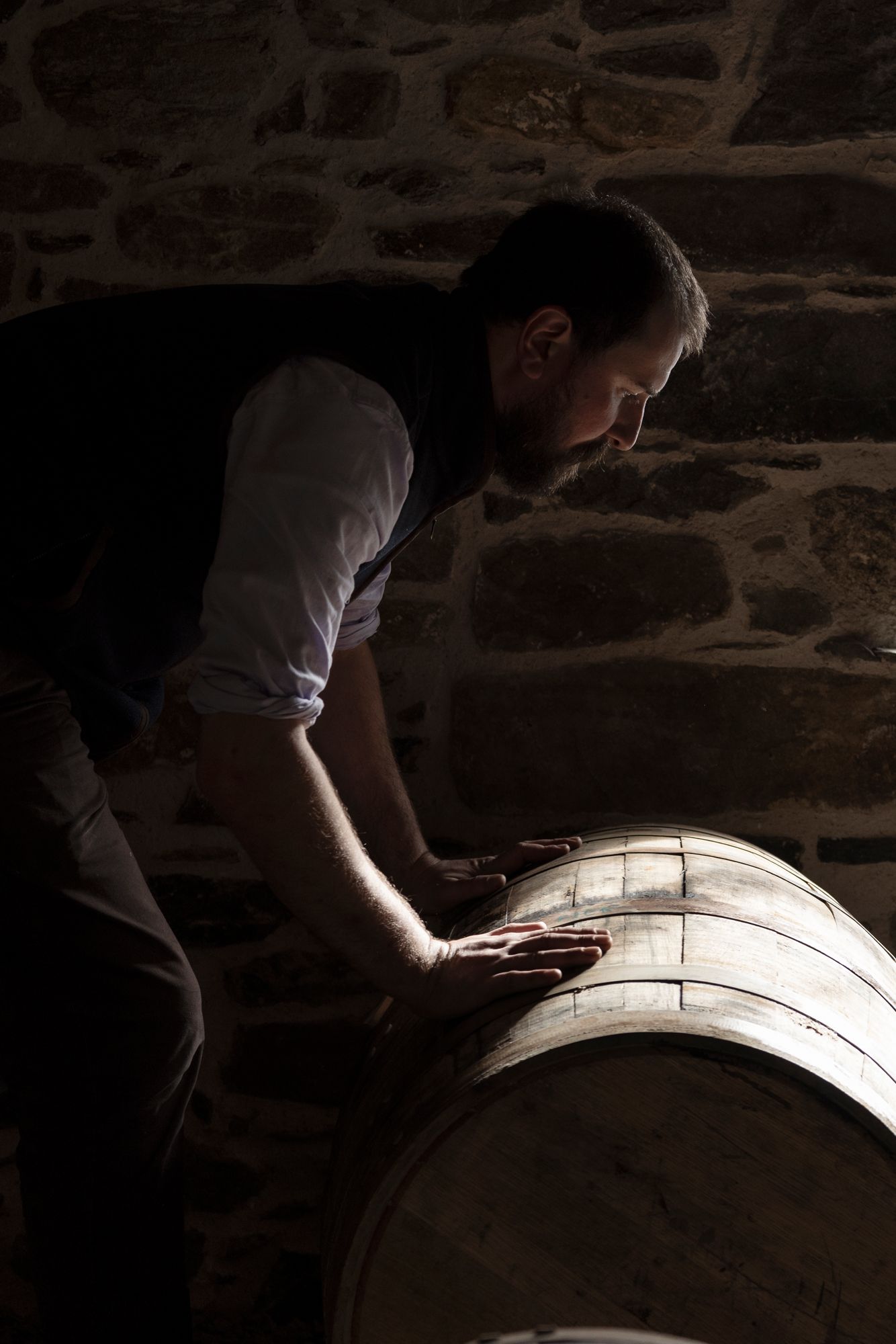 A person inspects a large whisky wooden barrel in a dimly lit cellar with stone walls, Scotland