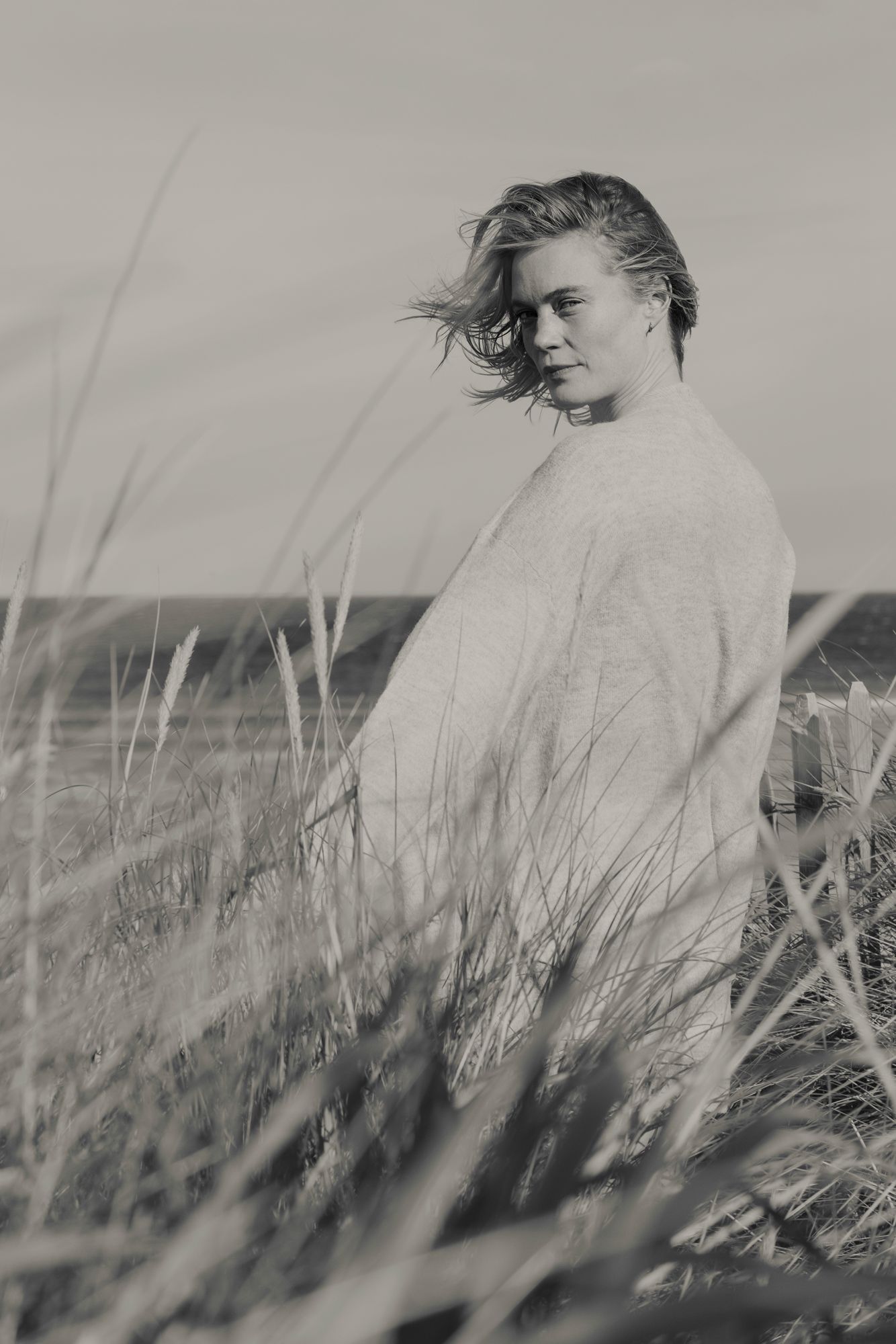 Black and white photograph of someone standing in tall grass on a windy day wearing a light coloured sweater. Photographer Susie Lowe