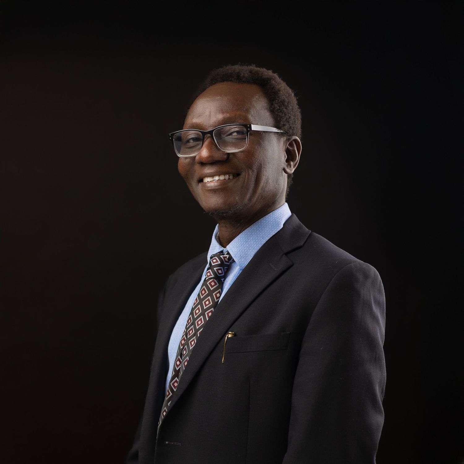 Professional headshot of someone wearing glasses and a suit with dotted tie against dark background, smiling warmly.
