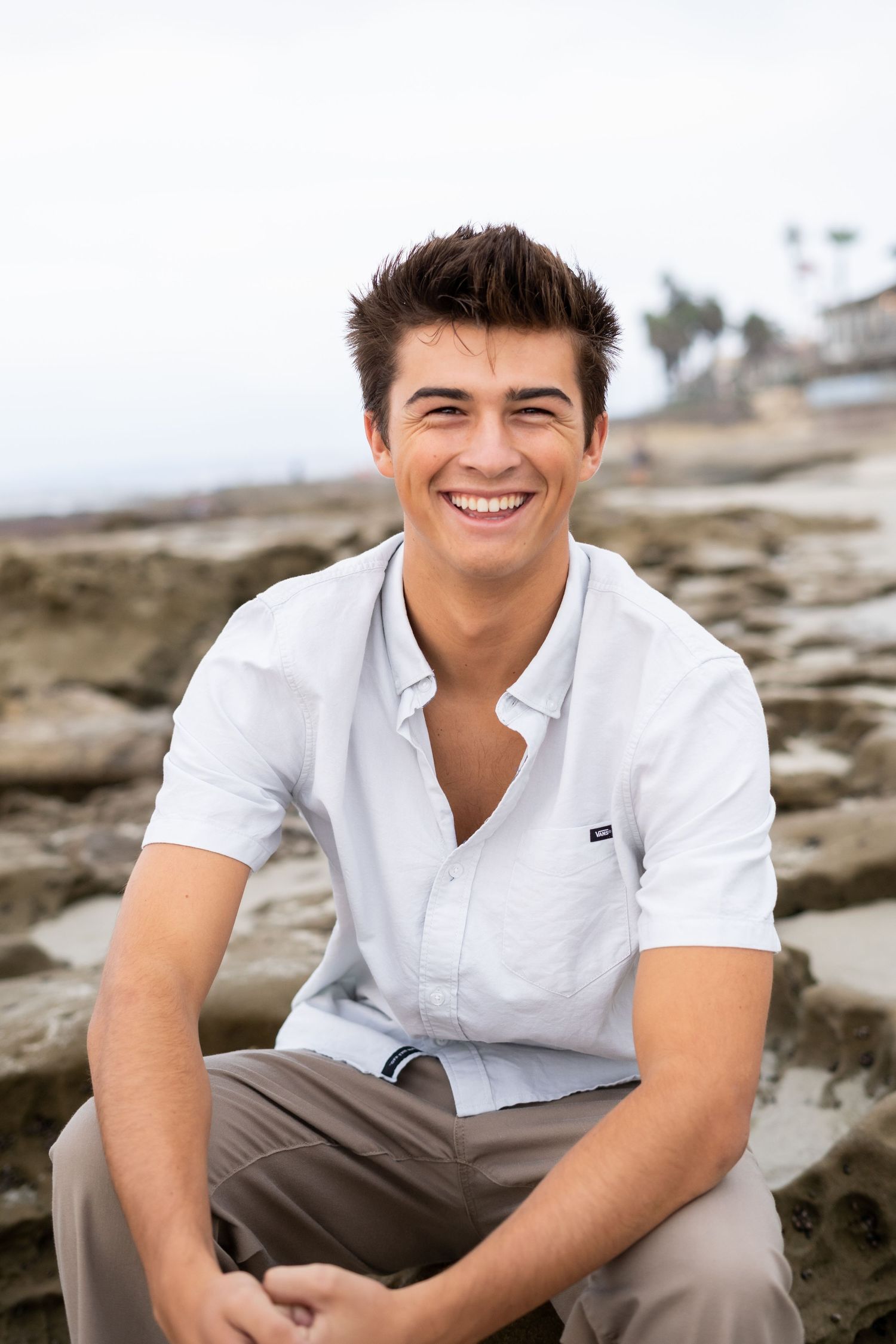 Portrait of a smiling high school senior wearing a white casual shirt sitting on coastal rocks at the beach.