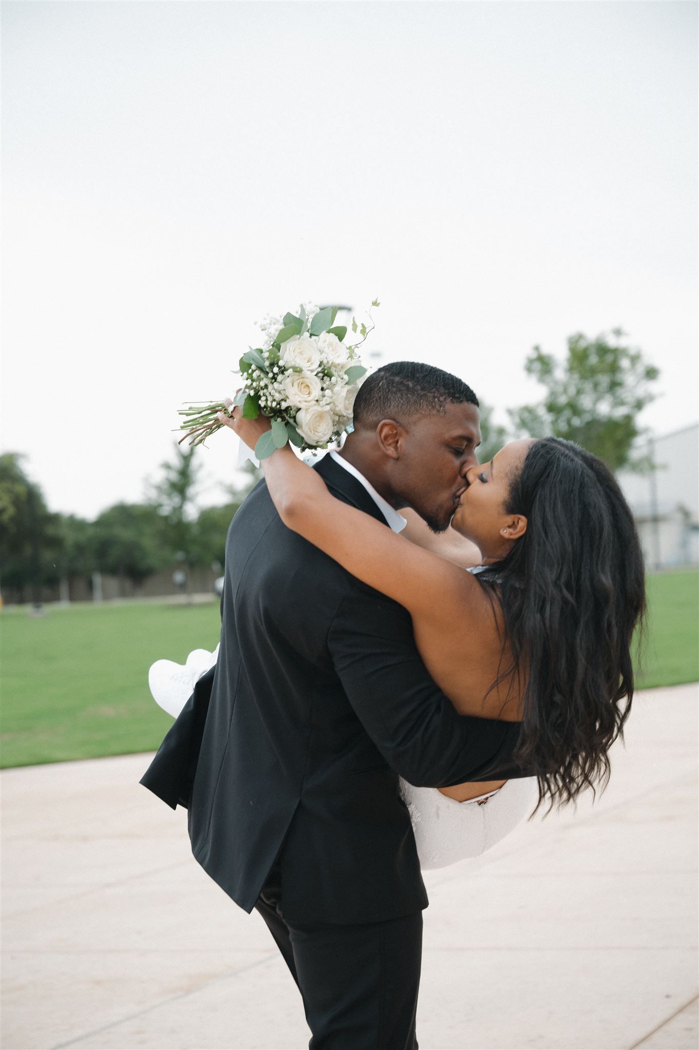 A romantic wedding couple shares a passionate kiss outdoors while holding a white floral bouquet.