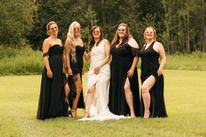 Bride in white dress stands with four bridesmaids wearing elegant black gowns outdoors.