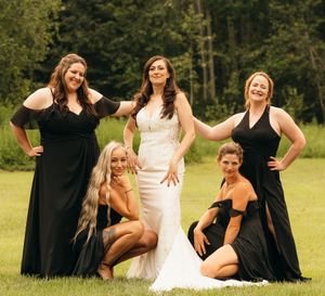 Bride in white gown poses with four bridesmaids in black dresses on green lawn.