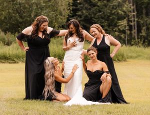 Bride in white gown surrounded by four bridesmaids in black dresses posing on a grassy field.