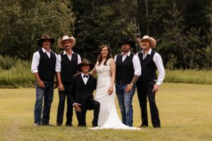 Bride in white gown poses with five groomsmen in black vests and cowboy hats on a green lawn.