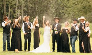 Wedding party celebrates with champagne spray outdoors wearing black attire and cowboy hats.