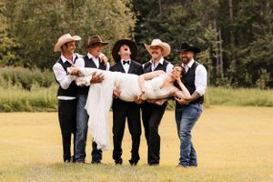Smiling groomsmen in cowboy hats and black vests carry the bride in a white gown outdoors.