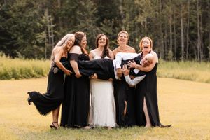 Four bridesmaids in black dresses playfully lift the groom while the bride stands smiling beside them.