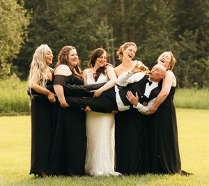 Bride in white gown laughing with four bridesmaids in black dresses lifting the groom outdoors.