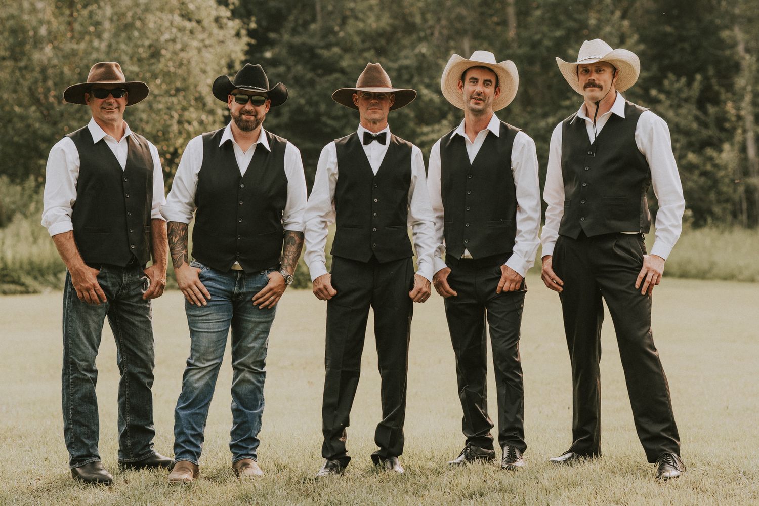 Five groomsmen in black vests and cowboy hats posing together on a green lawn outdoors.
