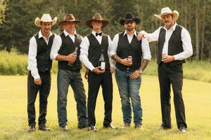 Five groomsmen in black vests and cowboy hats holding drinks while posing on a green lawn.