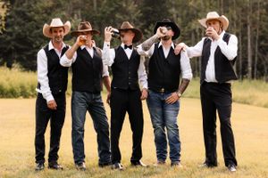 Groomsmen in cowboy hats and black vests toast drinks together on a sunny outdoor lawn.