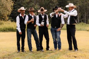 Five groomsmen in cowboy hats and black vests share a lighthearted moment on a green field.
