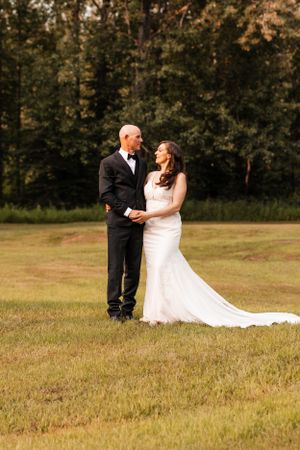 Wedding couple holding hands and gazing at each other in a sunlit grassy field with forest backdrop.