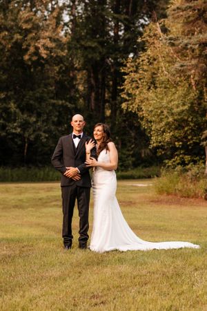 Smiling bride in white gown stands with groom in black tuxedo on grass with autumn trees behind.