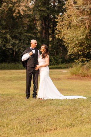 Bride in fitted white gown embraces groom in black suit in golden hour light at outdoor wedding.