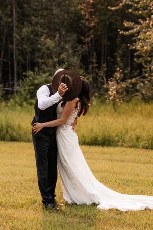 Bride and groom sharing a kiss hidden behind a brown cowboy hat in a green meadow setting.