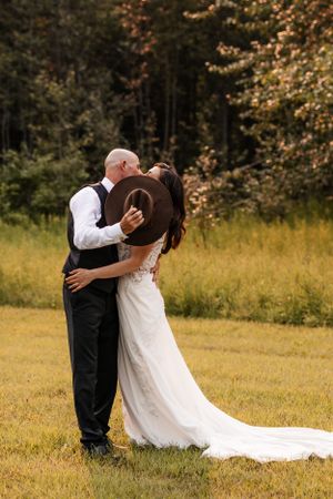 Wedding couple kissing behind a brown cowboy hat while embracing in a sunlit outdoor field.