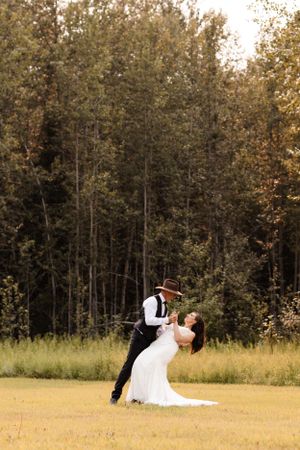 Groom dipping bride in a romantic dance pose in a grassy field surrounded by tall green trees.
