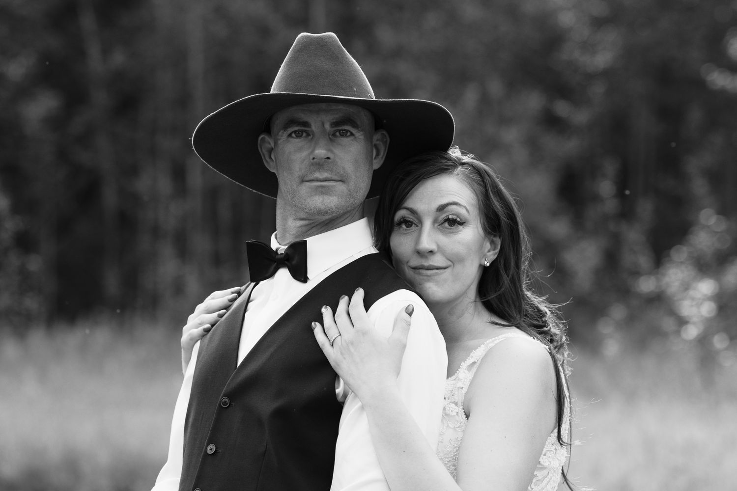 Black and white portrait of groom in cowboy hat and vest with bride leaning against him in a field.