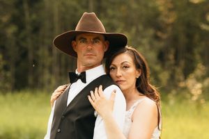Groom in brown cowboy hat and black vest standing with bride leaning against him in a sunny field.