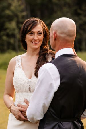 Radiant bride in lace wedding gown smiling while holding hands with groom in black vest outdoors.