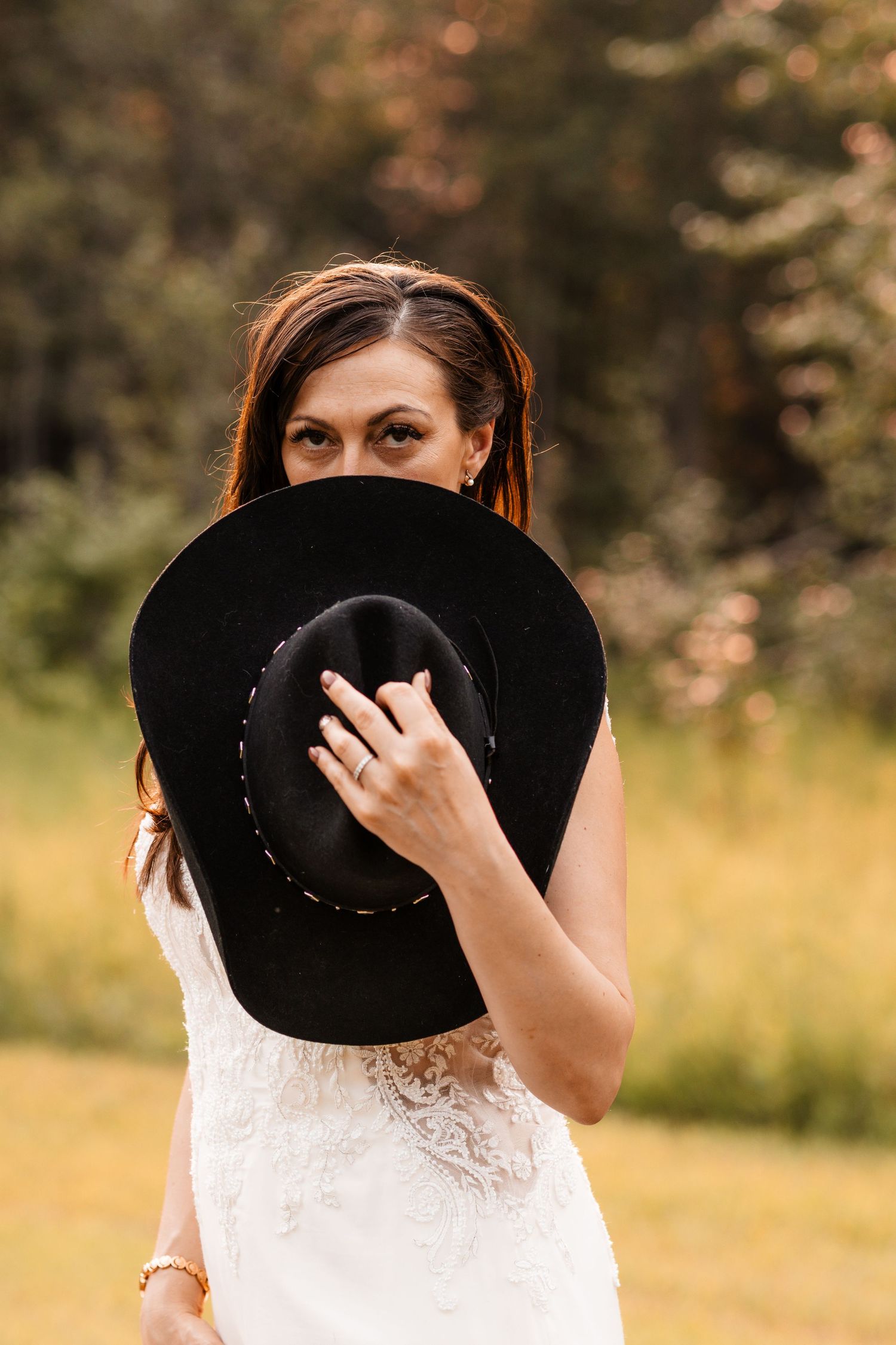Bride in lace wedding dress playfully hides behind a black wide-brim hat in a green field.