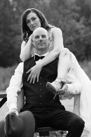 Black and white portrait of bride draped over seated bald groom holding a cowboy hat outdoors.
