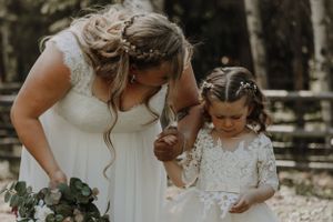 Bride in white lace gown bends toward young flower girl in a matching white dress outdoors.