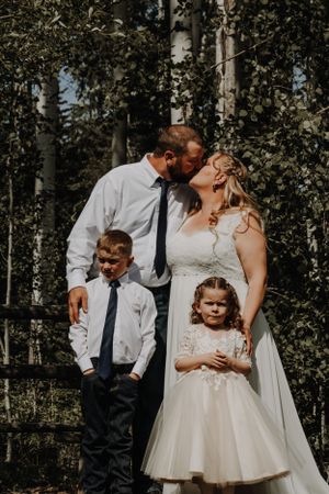 Bride and groom kiss while two children stand beside them in front of a birch tree forest.