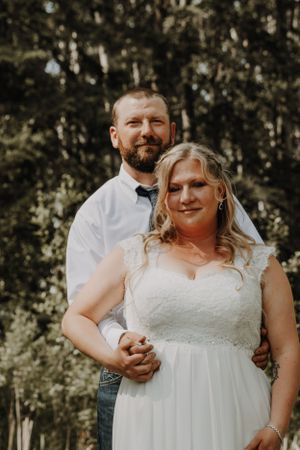 Bride and groom pose together in a forest, with groom standing behind bride in a white wedding gown.