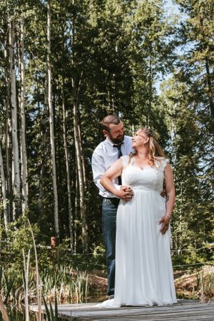 Bride in white gown and groom share a joyful moment on a wooden deck surrounded by tall trees.