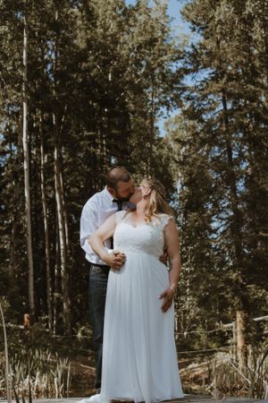 Bride and groom share a romantic kiss outdoors surrounded by tall evergreen trees on their wedding day.