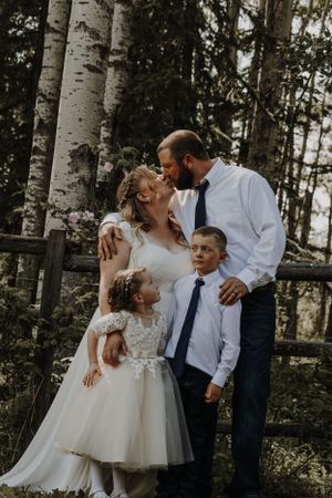 Bride and groom share a kiss while their two children stand close in a lush forest setting.