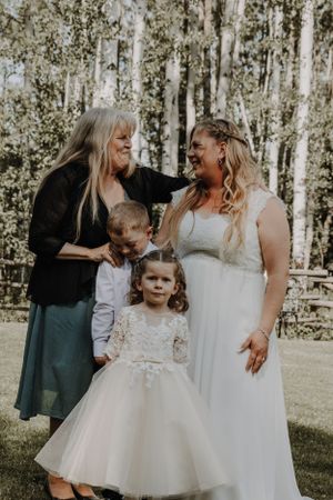 Bride in white gown laughs with older woman and two children in a sunlit forest setting.
