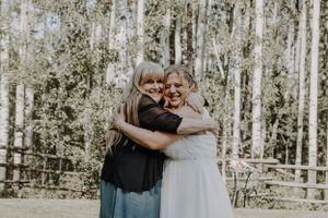 Bride in white dress sharing warm hug with older woman at outdoor wedding.