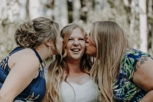 Bride laughing as two bridesmaids in floral dresses kiss her cheeks outdoors.