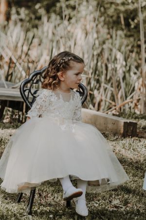 Young flower girl in white lace tulle dress seated on chair outdoors.