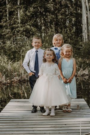 Four children posing on wooden dock in wedding attire near forest pond.