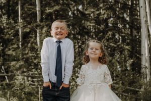 Boy in navy tie and girl in white flower girl dress smiling outdoors.