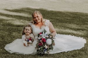 Smiling bride holding bouquet seated on lawn with flower girl in white dress.