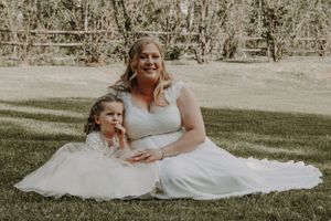 Bride in white gown sitting on grass with young flower girl.
