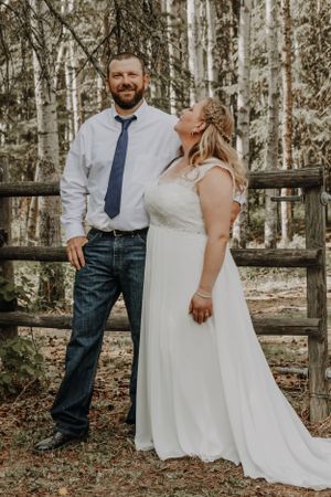 Smiling groom in blue tie stands with bride in white lace gown by wooden fence.