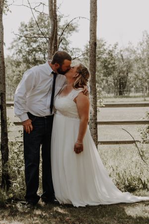Bride and groom share a romantic kiss near rustic wooden fence surrounded by trees.