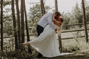 Groom dips bride in white flowing gown for a romantic kiss among birch trees.