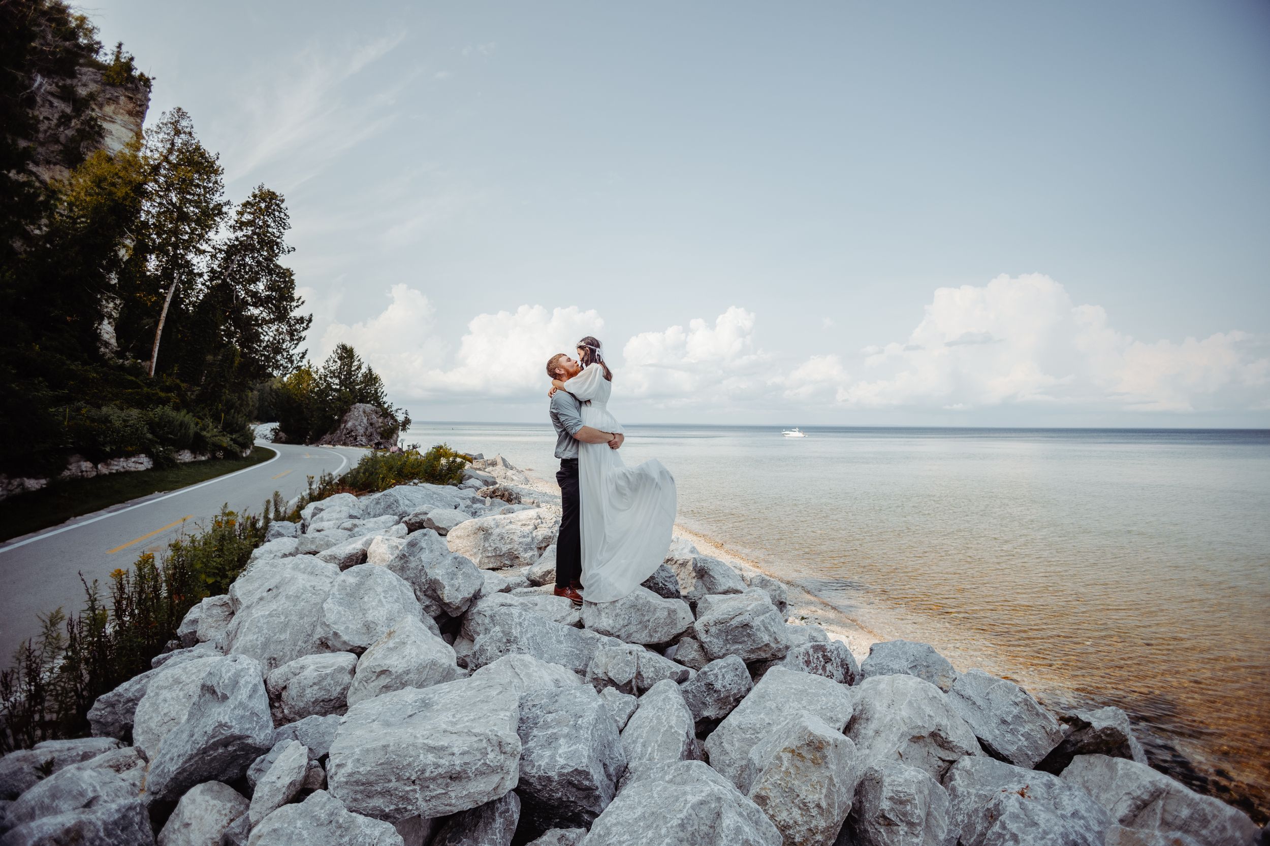 Newlyweds sharing intimate moment on rocky shoreline overlooking serene water.